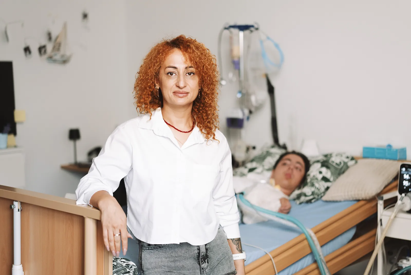 Angehörige steht am Pflegebett eines beatmeten jungen Mannes, im Hintergrund das Beatmungsgerät