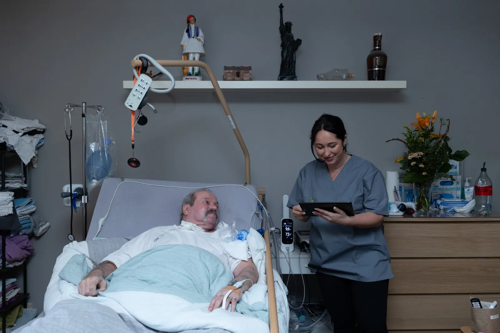 Pflegerin dokumentiert am Tablet am Bett eines Mannes, häuslich eingerichtetes Zimmer mit Andenken im Regal