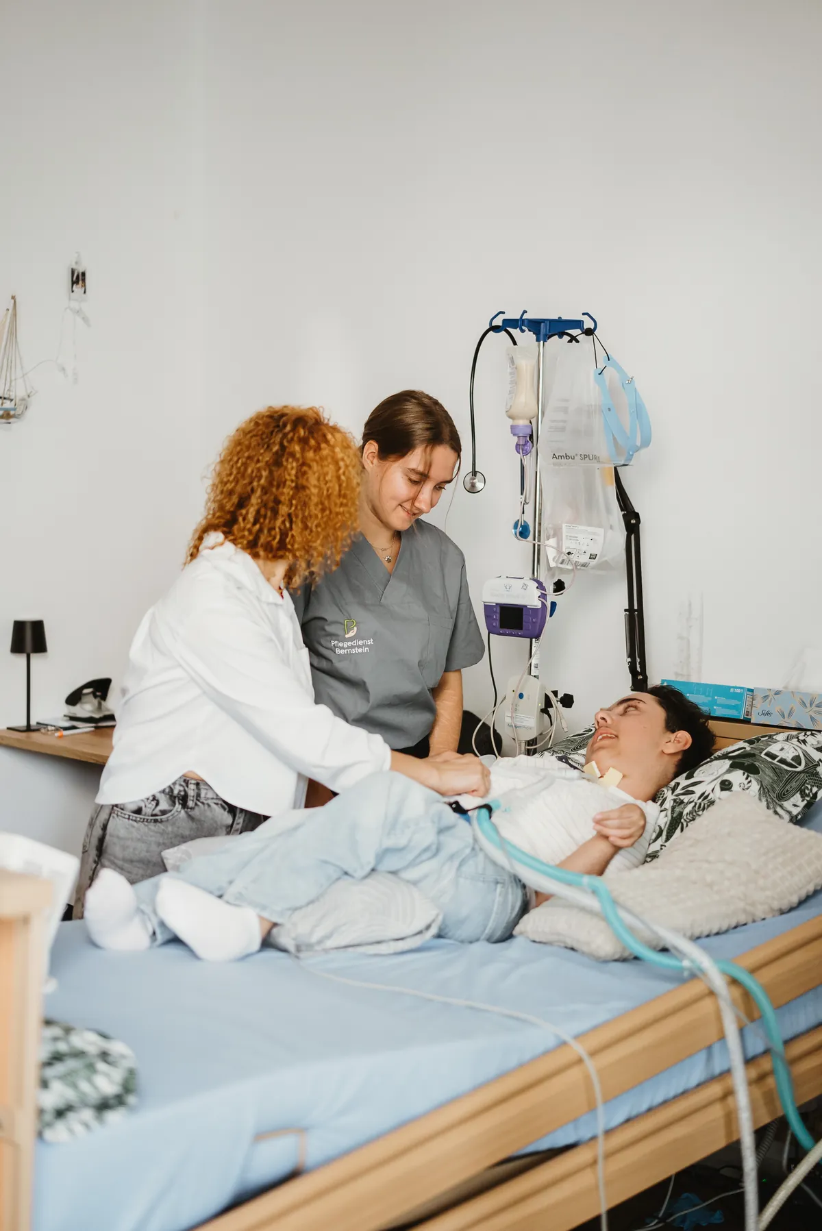 Angehörige und Pflegerin im Bernstein-Kasack am Bett eines beatmeten Patienten, Beatmungsschlauch und Infusionsständer daneben