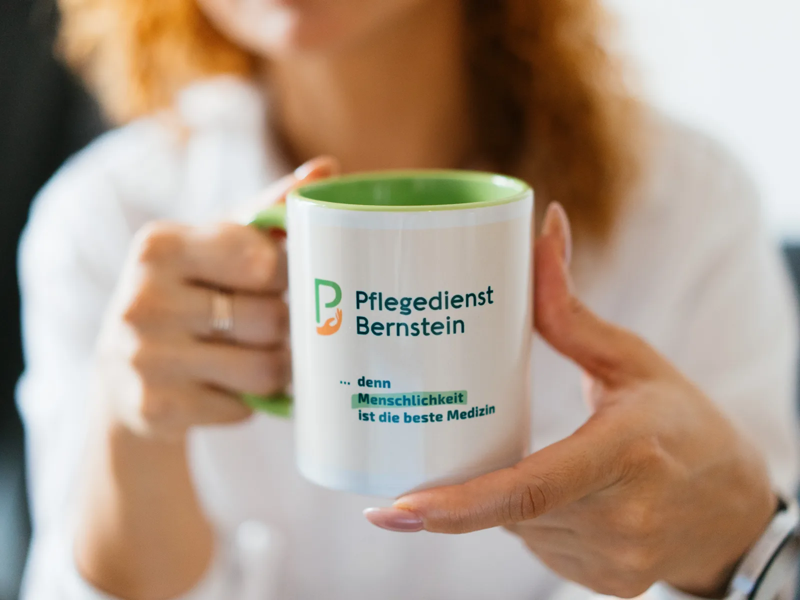 Frau in weißer Bluse hält eine weiße Tasse mit grünem Innenrand, darauf das Logo des Pflegedienst Bernstein und der Satz denn Menschlichkeit ist die beste Medizin