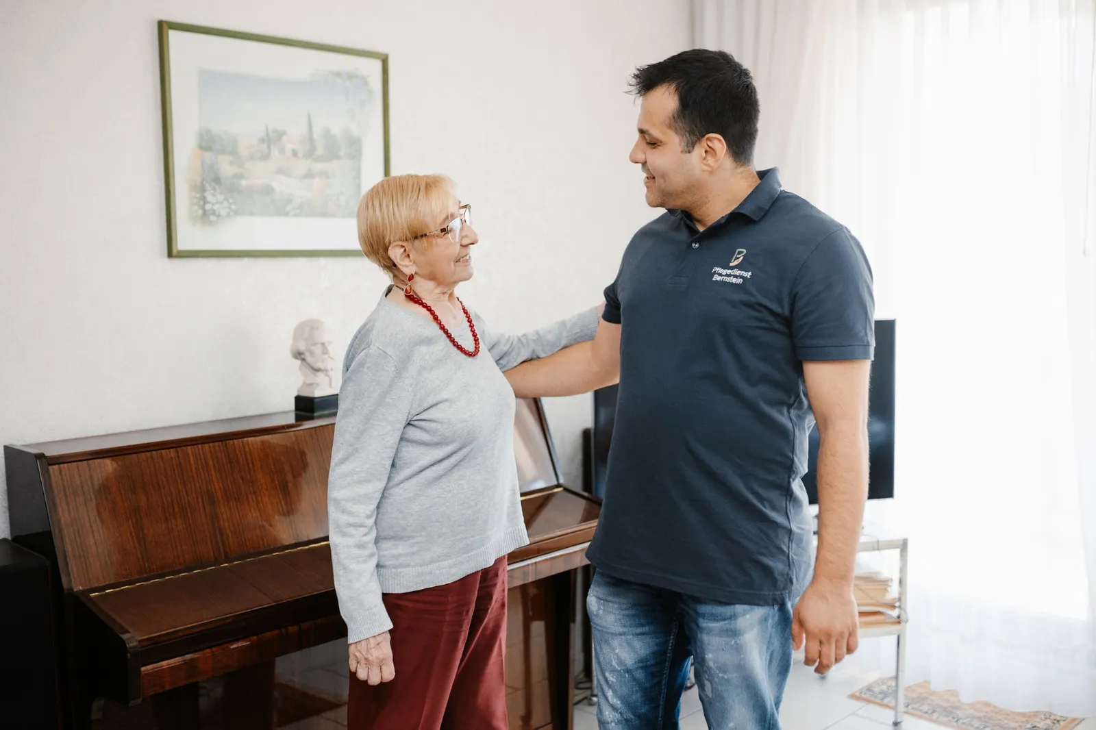 Pfleger und ältere Dame stehen einander zugewandt in einem Wohnzimmer neben einem Klavier