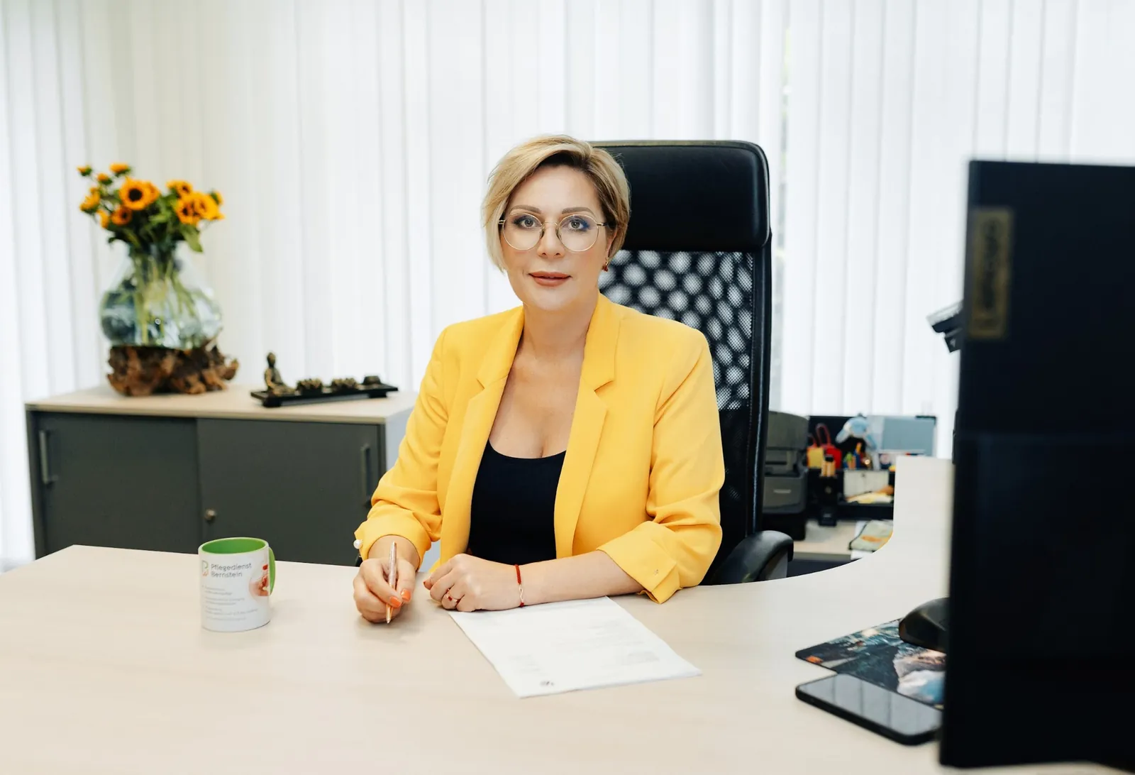 Geschäftsführerin Svetlana Petelin im gelben Blazer an ihrem Schreibtisch, daneben eine Bernstein-Tasse und Sonnenblumen