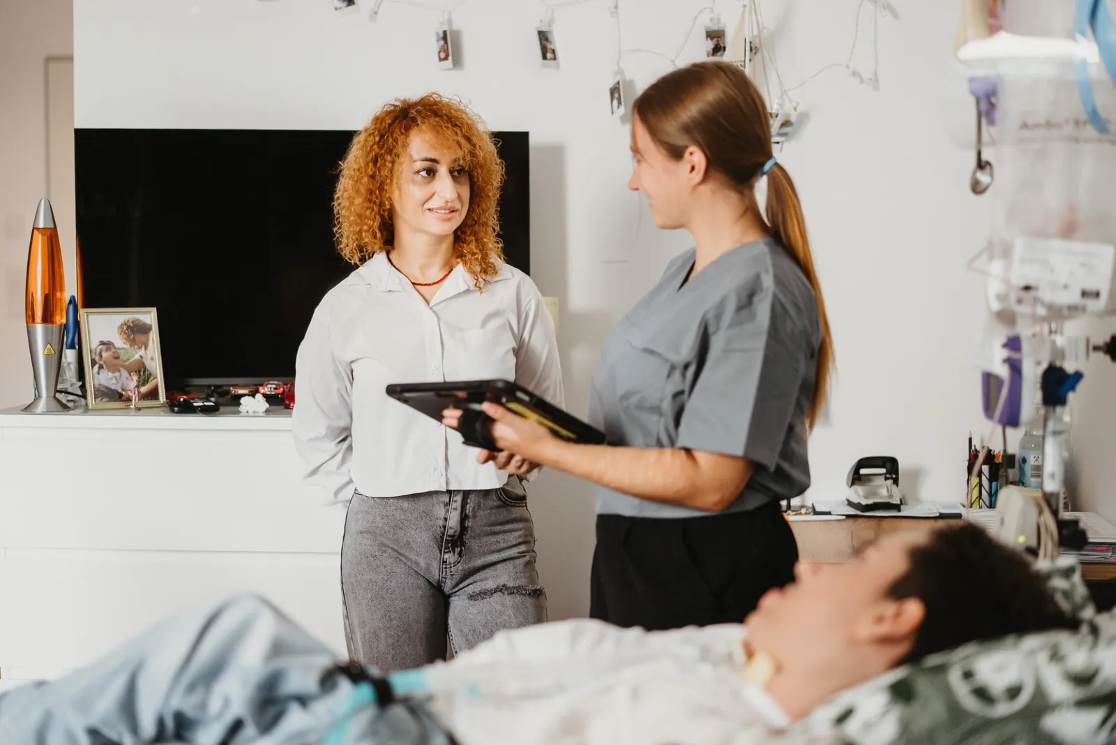 Pflegerin im Bernstein-Kasack bespricht am Tablet etwas mit einer Angehörigen, im Vordergrund ein Patient im Pflegebett, daneben Beatmungs- und Infusionstechnik