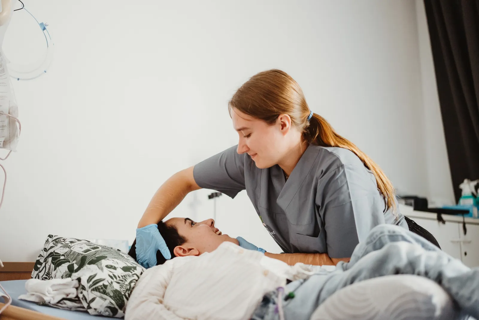 Pflegefachkraft mit Handschuhen streicht einem Patienten im Pflegebett über den Kopf, links ein Infusionsständer