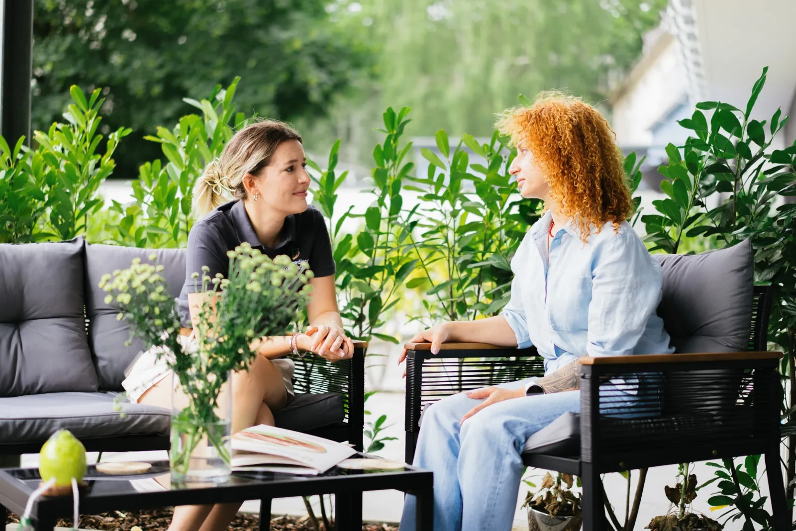 Zwei Frauen im Gespräch auf einer begrünten Terrasse, Unterlagen und Getränke auf dem Tisch