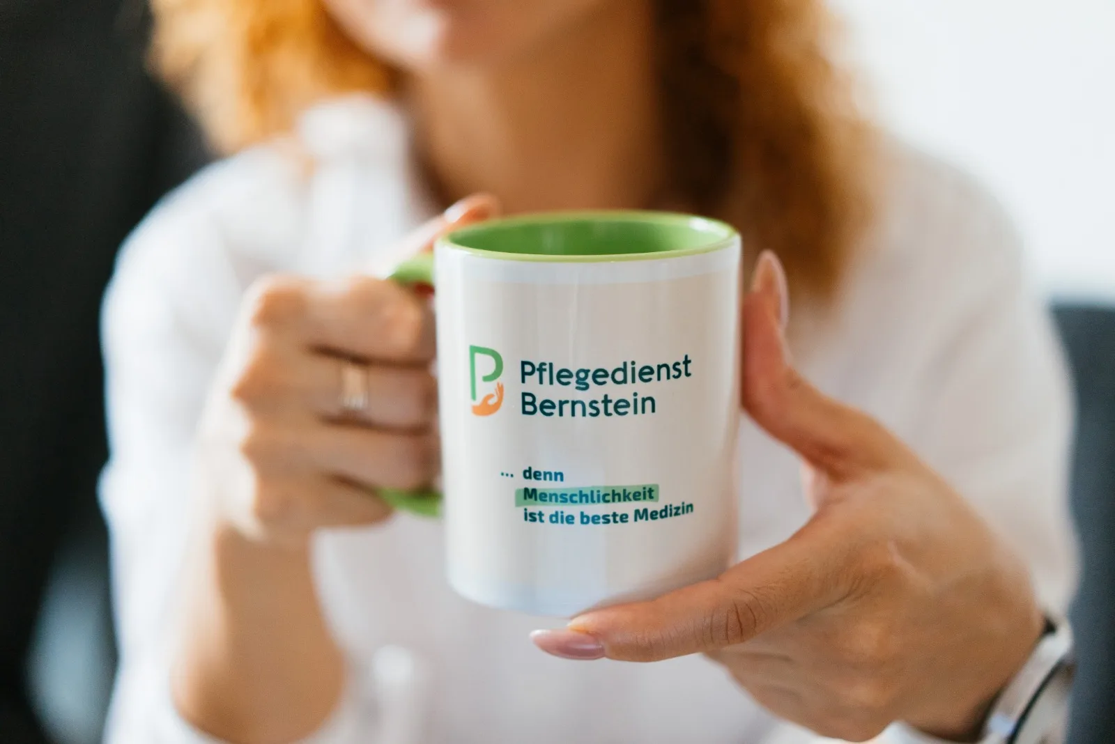 Hände halten eine Tasse mit dem Logo und dem Slogan des Pflegedienst Bernstein: denn Menschlichkeit ist die beste Medizin