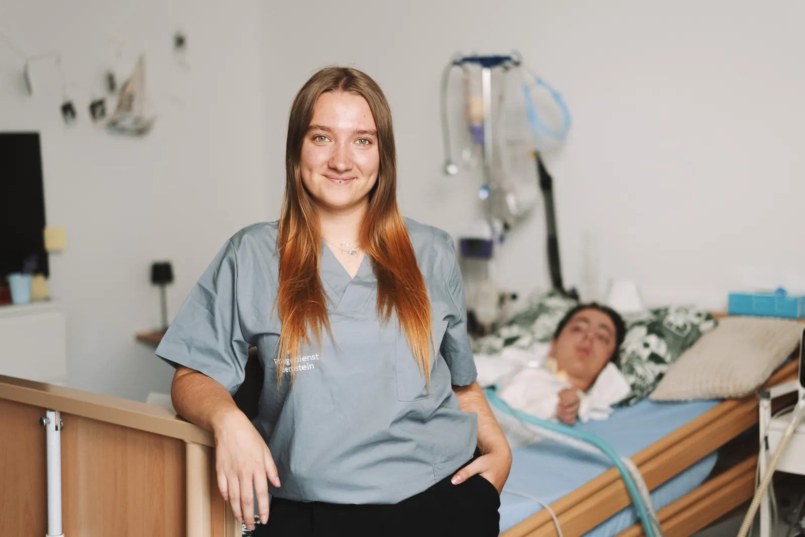 Junge Pflegefachkraft im grauen Bernstein-Kasack steht lächelnd am Pflegebett, dahinter ein Patient und ein Beatmungsgerät