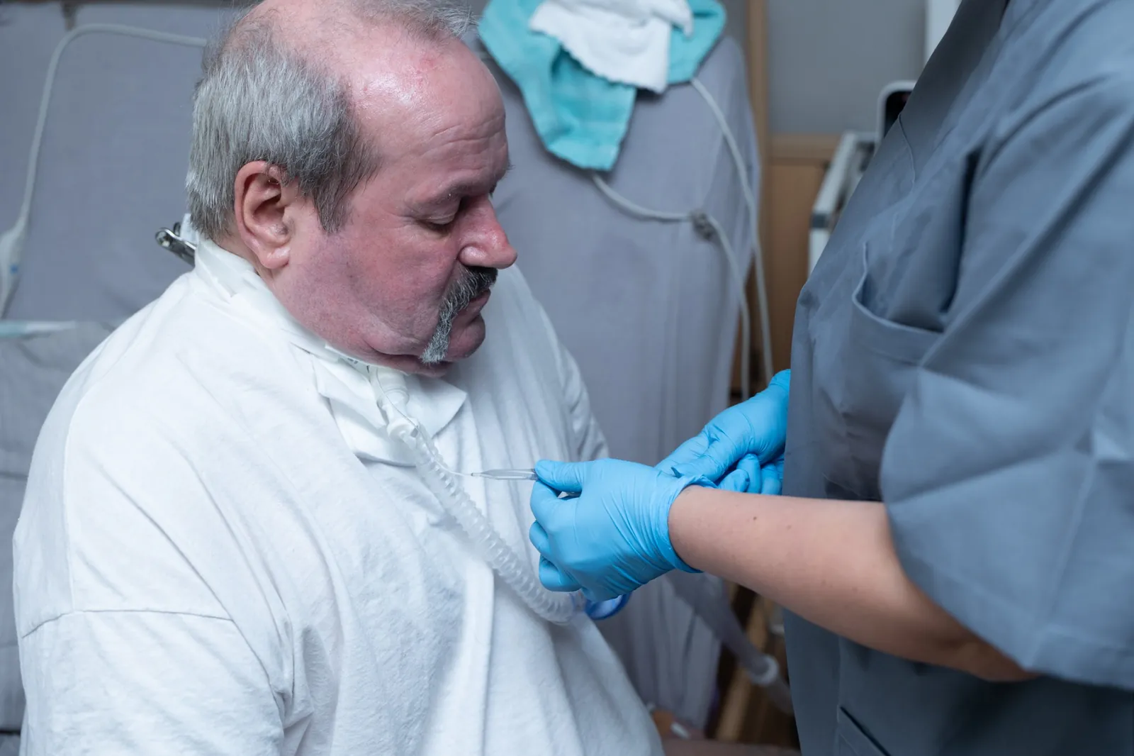 Pflegerin mit Handschuhen versorgt den Kanülenanschluss am Hals eines älteren Mannes, Halteband und Schlauch sind zu sehen